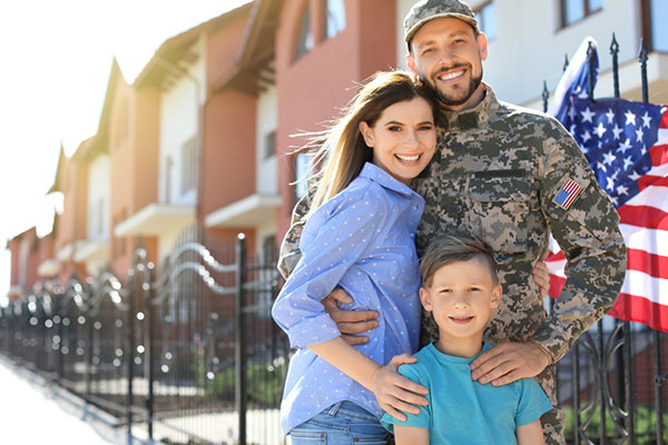 A man in Army fatigues stands in front of a home with a woman and a young boy.