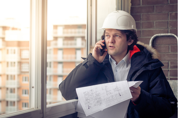 contractor in winter coat speaks on a phone standing next to a window
