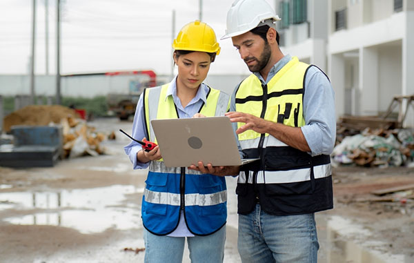 Two people look at a computer on a construction site.