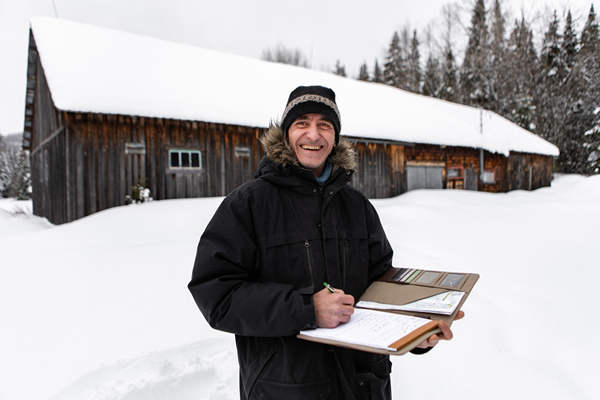 Smiling home inspector with a clipboard in front of a snowy barn.