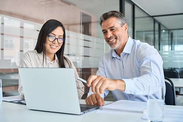 A woman with long black hair and a man with gray hair sit at a workstation in front of a laptop.