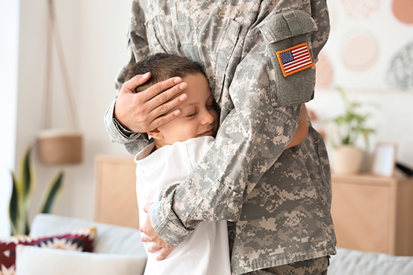 A female soldier in U.S. fatigues hugs a young boy while standing in a living room.