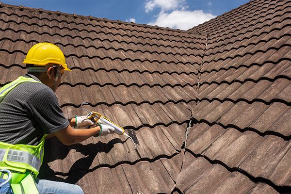 A man wearing a yellow safety vest works to seal a crack on a tile roof.