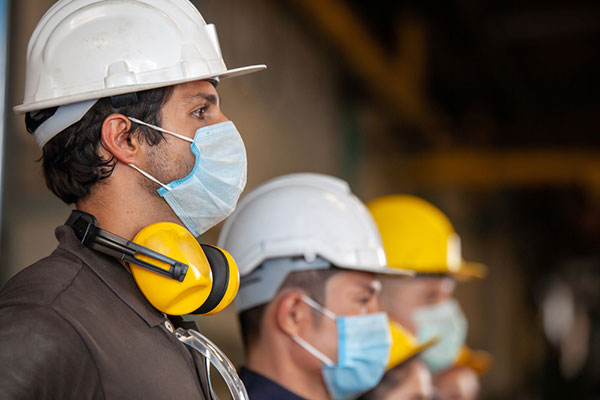 Construction workers wearing masks, hard hats, and ear protection wait in line for instructions
