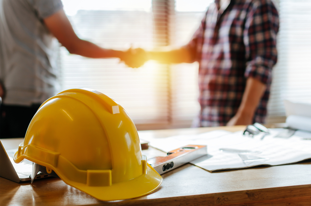 Contractor shaking hands with a new client in the background with a hard hat on desk in foreground