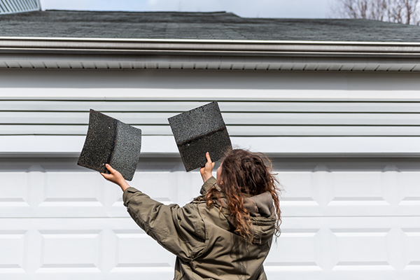A woman looks at a roof holding damaged shingles.